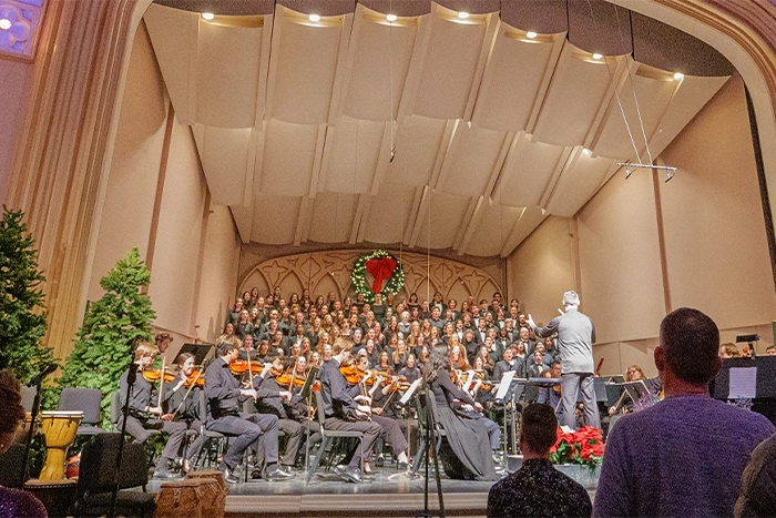 An auditorium with a stage full with an orchestra, chorus, and holiday greenery